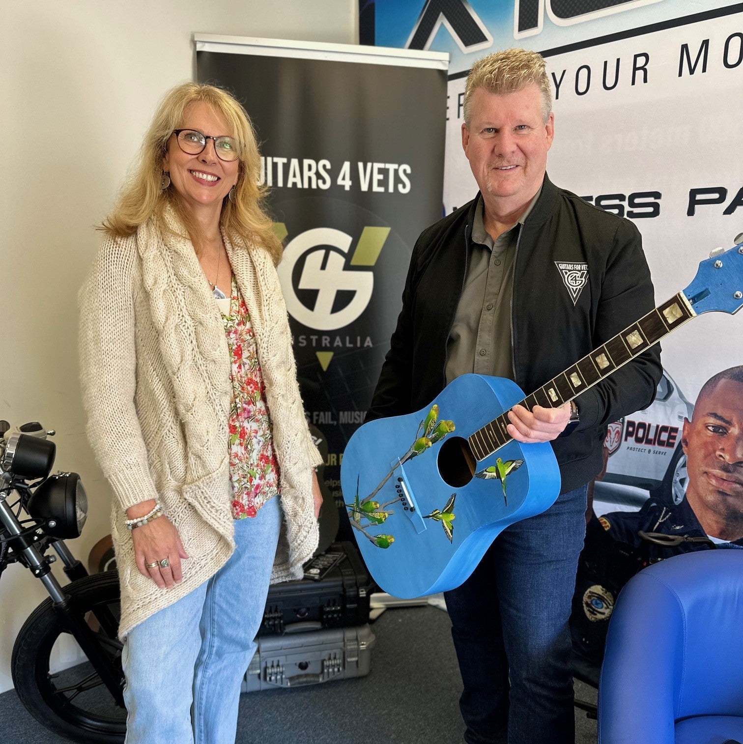 Man holding a blue guitar next to a woman in an indoor setting with promotional banners in the background.